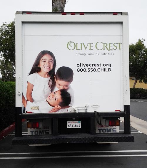 Back of a white box truck showing a girl and her two brothers huddled and smiling, all wearing white shirts. Also shown are a non-profit organization's logo and contact information.