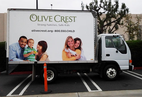 Side of a white box truck showing two sets of families. On the left is a multi-cultural family with a father in a blue shirt, a young boy in a green shirt and a mother in a light-colored blouse. The child is being held by his parents on either side. On the right of the truck is a blonde mother in yellow, shown with her teen daughter, who resembles her. Also shown are a non-profit organization's logo and contact information.