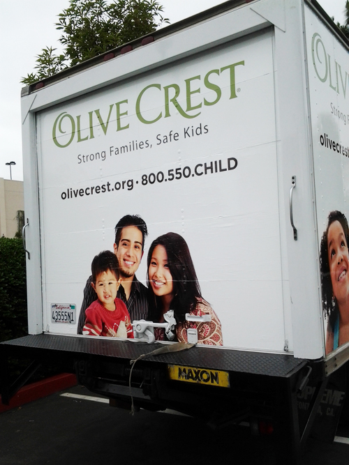 The back of a second white box truck shows a happy family with a father, mother and little boy. The little boy is wearing a red shirt, while the father is wearing a dark shirt and the mother is wearing a pinkish patterned blouse. Also shown are a non-profit organization's logo and contact information.