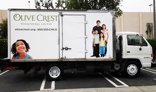 The side of a second white box truck shows a family of four standing together: mother, father, brother and sister, all smiling at the camera. Their photo appears beside a door on the truck that goes into the cargo area. On the other side of the door is a young girl grinning and looking upward at a logo and organization's contact info.
